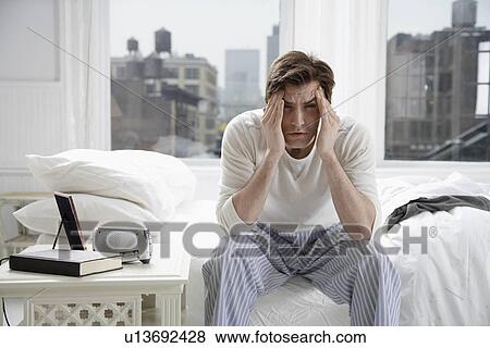 Young man sitting on bed and rubbing forehead View Large Photo Image Stock Photo - Young man sitting on bed and rubbing forehead. Fotosearch
