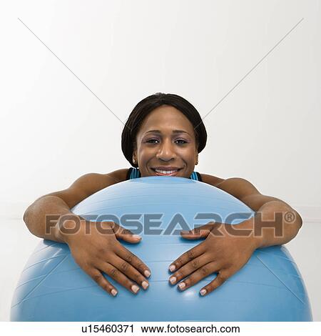 Stock Image - African American young adult woman resting with arms draped over exercise ball smiling at viewer.. Fotosearch