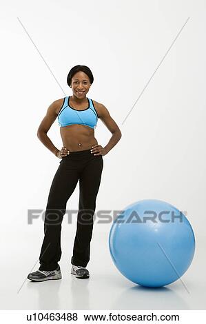 Stock Photo - African American young adult woman smiling at viewer with hands on hips next to exercise ball.. Fotosearch