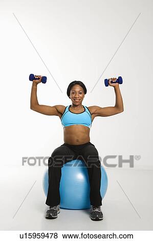 African American young adult woman sitting on exercise ball and raising dumbbells over head. View Large Photo Image Stock Photo - African American young adult woman sitting on exercise ball and raising dumbbells over head.. Fotosearch