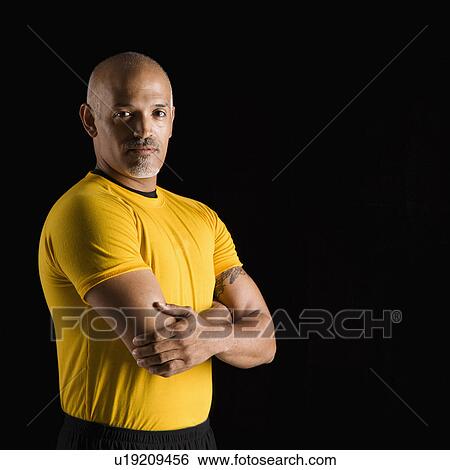 Stock Photograph - Mid adult multiethnic man wearing yellow exercise shirt with arms crossed looking at viewer.. Fotosearch