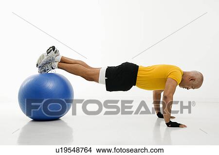 Picture - Side view of mid adult multiethnic man doing pushups while balancing on blue exercise ball.. Fotosearch