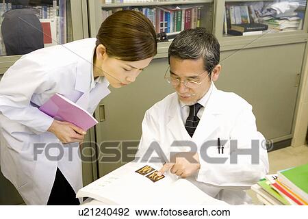 Stock Image - Mature scientist man explaining to a young scientist woman in laboratory, side view, high angle view. Fotosearch