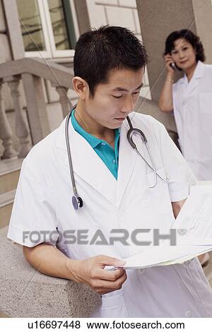 Close-up of a male doctor reading a report with a female doctor talking on mobile phone in the background View Large Photo Image Stock Photo - Close-up of a male doctor reading a report with a female doctor talking on mobile phone in the background. Fotosearch
