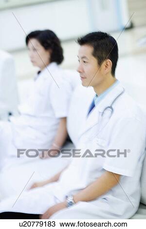 Stock Image - Side profile of a male doctor sitting on a couch with a female doctor sitting behind him. Fotosearch