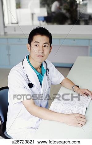 Stock Image - Side profile of a male doctor sitting at the table and holding a book. Fotosearch