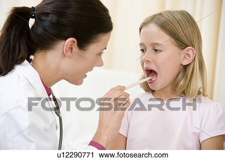 Doctor giving checkup to young girl with tongue depressor in exam room View Large Photo Image Stock Image - Doctor giving checkup to young girl with tongue depressor in exam room. Fotosearch