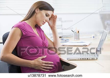 Stock Image - Pregnant woman at work with laptop looking stressed. Fotosearch