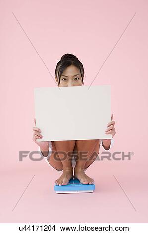 Woman crouching on bathroom scale holding blank placard View Large Photo Image Picture - Woman crouching on bathroom scale holding blank placard. Fotosearch