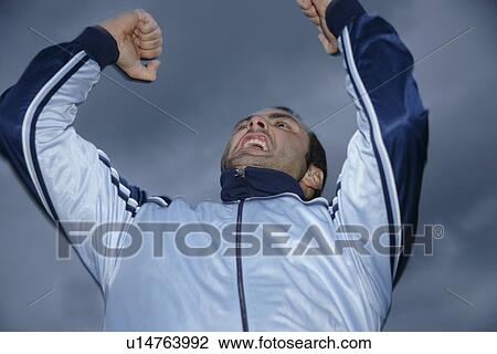 Stock Image - Young man in a sweat suit jacket cheering in front of dark clouds. Fotosearch
