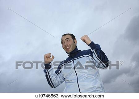 Stock Image - Young man in a sweat suit jacket cheering in front of dark clouds. Fotosearch