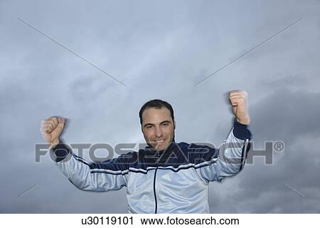 Stock Image - Young man in a sweat suit jacket cheering in front of dark clouds. Fotosearch