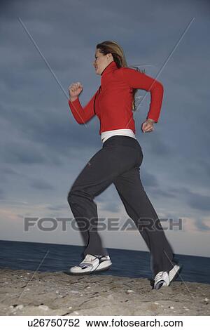 Stock Image - Young woman jogging at the beach, full length, side view. Fotosearch