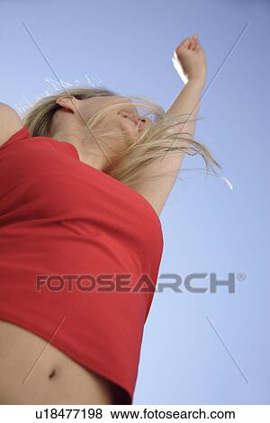 Stock Photo - Young woman dancing and cheering with arms stretched out in front of blue sky, low angle view. Fotosearch