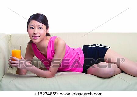Stock Photo - Young woman holding a glass of orange juice and lying on sofa. Fotosearch