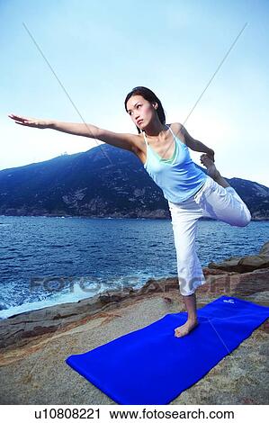 Stock Image - Young woman practising yoga on exercise mat, near the sea. Fotosearch
