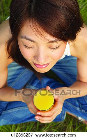 Stock Photo - Young woman sitting on grass, eyes closed, holding glass of drink. Fotosearch