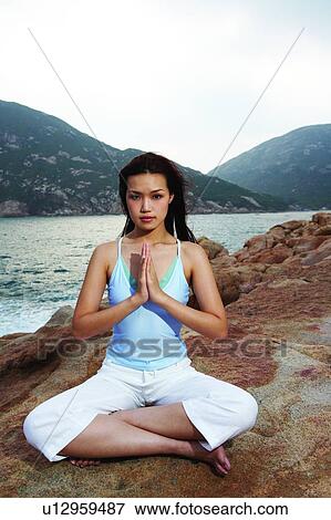 Stock Photo - Young woman sitting on rock, near the sea, in Sukasana position. Fotosearch