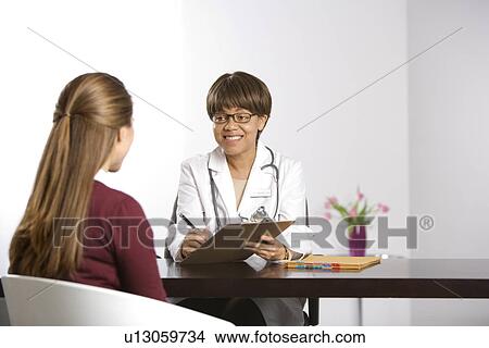 African American middle-aged female doctor sitting at desk talking to Caucasian mid-adult female patient taking notes on clipboard. View Large Photo Image Picture - African American middle-aged female doctor sitting at desk talking to Caucasian mid-adult female patient taking notes on clipboard.. Fotosearch