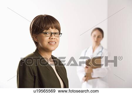 Stock Photo - African American middle-aged female patient woman smiling looking at viewer with Caucasian mid-adult female doctor standing in background.. Fotosearch