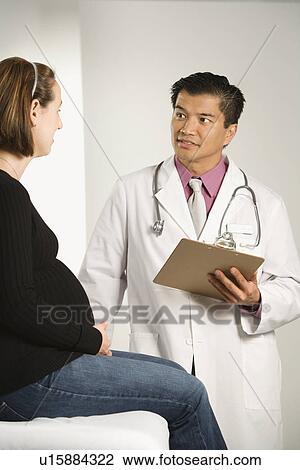 Stock Image - Asian American male doctor examining Caucasian pregnant female patient.. Fotosearch