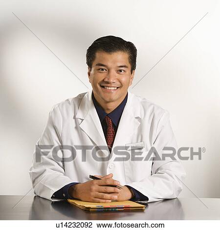 Stock Image - Asian American male doctor sitting at desk with charts smiling and looking at viewer.. Fotosearch