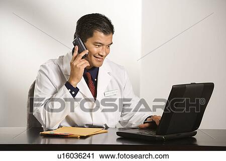 Stock Image - Asian American male doctor sitting at desk with charts and laptop computer talking on cellphone.. Fotosearch
