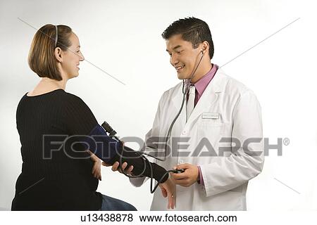 Stock Photo - Asian American male doctor testing blood pressure of pregnant Caucasian woman.. Fotosearch