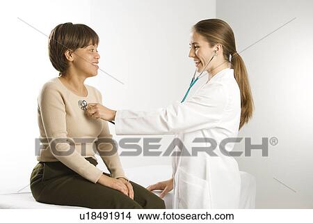 Picture - Caucasian mid-adult female doctor examining African American middle-aged female patient with stethoscope.. Fotosearch