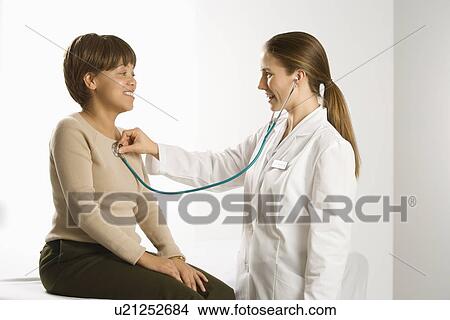 Caucasian mid-adult female doctor examining African American middle-aged female patient with stethoscope. View Large Photo Image Picture - Caucasian mid-adult female doctor examining African American middle-aged female patient with stethoscope.. Fotosearch