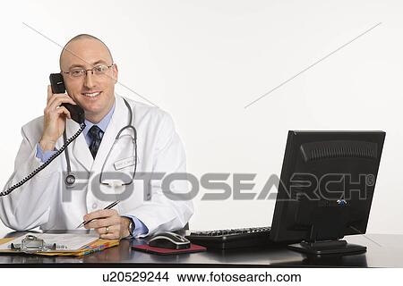 Picture - Caucasian mid adult male physician sitting at desk with laptop computer.. Fotosearch