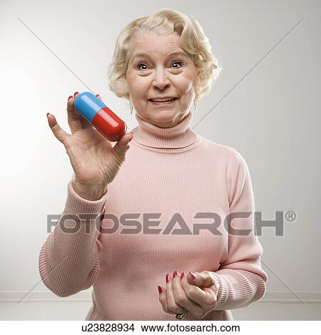 Picture - Caucasian senior woman holding oversized pill at viewer.. Fotosearch