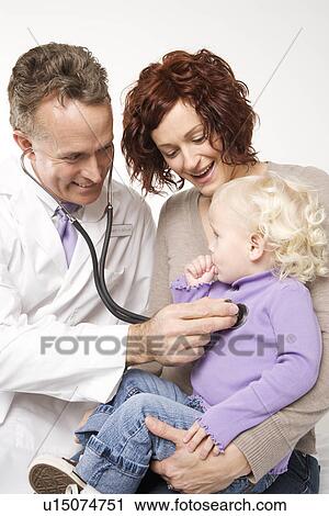 Stock Image - Middle-aged adult Caucasian male doctor holding stethoscope to female toddler's chest with mother watching.. Fotosearch