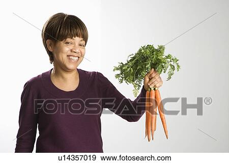 Stock Photo - Portrait of African American middle-aged woman holding bunch of fresh carrots smiling and looking at viewer.. Fotosearch