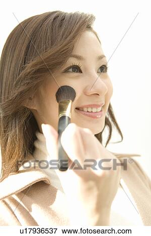 Stock Photo - A woman applying blusher on cheek, smiling, side view, white background, low angle view. Fotosearch