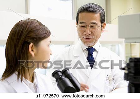 Stock Image - Male scientist talking to female scientist sitting in front of a microscope, front view, side view. Fotosearch