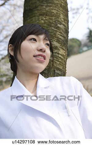 Stock Photo - Woman in lab coat leaning on a tree trunk, front view, Japan. Fotosearch