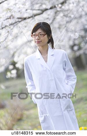 Stock Photography - Woman in lab coat standing under the cherry tree, front view, Japan. Fotosearch