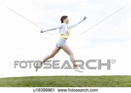 Stock Image - Young Woman jumping on lawn, Arms Outstretched, low angle view. Fotosearch