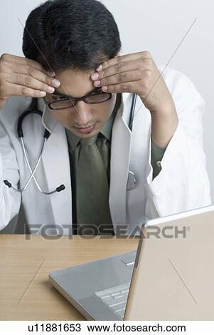 Stock Image - Close-up of a male doctor sitting in front of a laptop looking stressed. Fotosearch