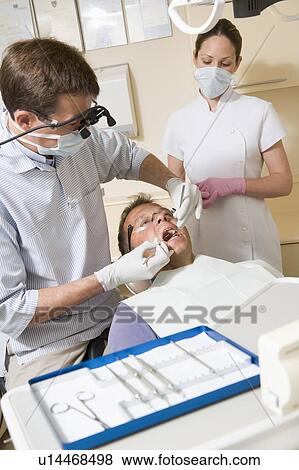 Stock Photo - Dentist and assistant in exam room with man in chair. Fotosearch