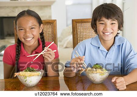 Picture - Two young children eating Chinese food in dining room smiling. Fotosearch