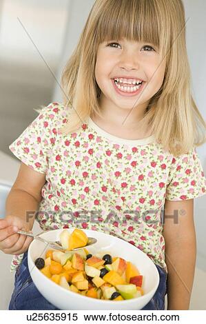 Stock Photography - Young girl in kitchen eating bowl of fruit smiling. Fotosearch