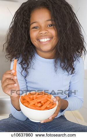 Stock Image - Young girl in kitchen eating carrot sticks smiling. Fotosearch