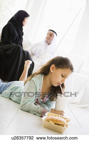 Stock Image - Young girl with fast food in living room with parents in background (high key/selective focus). Fotosearch