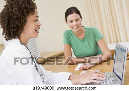 Stock Image - Doctor using laptop with woman in doctor's office smiling. Fotosearch
