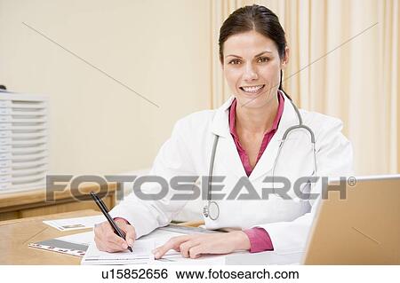 Stock Photograph - Doctor with laptop writing in doctor's office smiling. Fotosearch