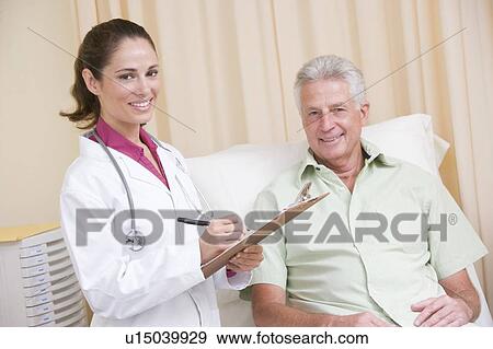 Stock Photo - Doctor writing on clipboard while giving checkup to man in exam room smiling. Fotosearch