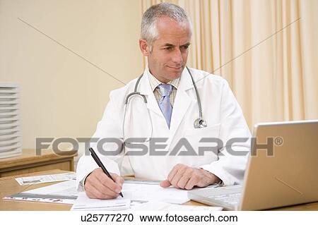 Stock Image - Doctor using laptop and writing in doctor's office. Fotosearch