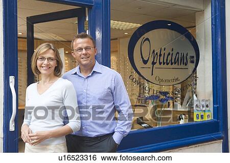 Stock Photograph - Couple standing at front entrance of optometrists smiling. Fotosearch